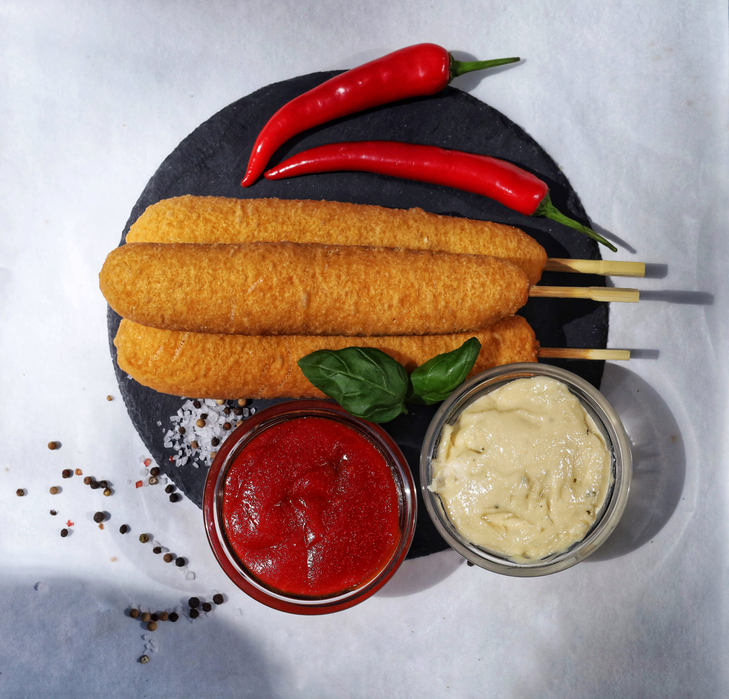 Corn dogs with sauces and red chili peppers on a black plate